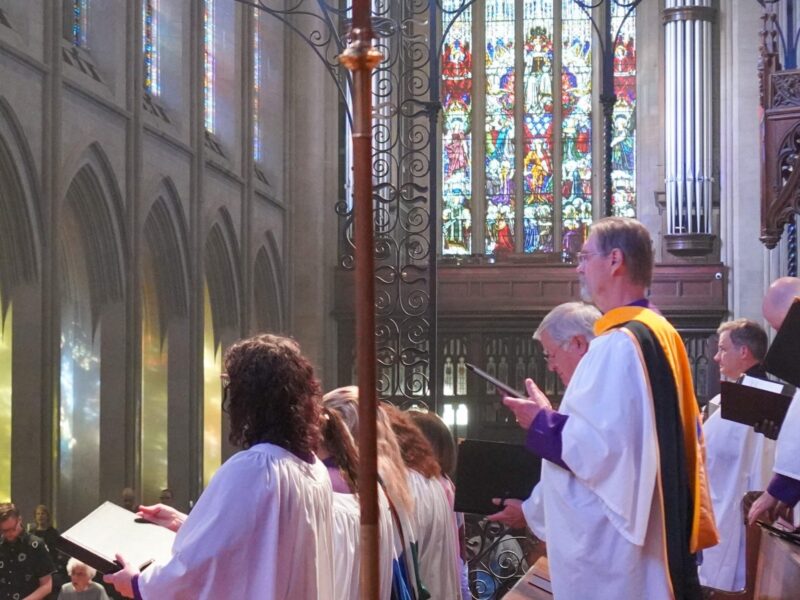Choir members in white robes sing during Choral Evensong at St. John’s Cathedral in Denver, with stained glass windows casting colorful light across the nave.