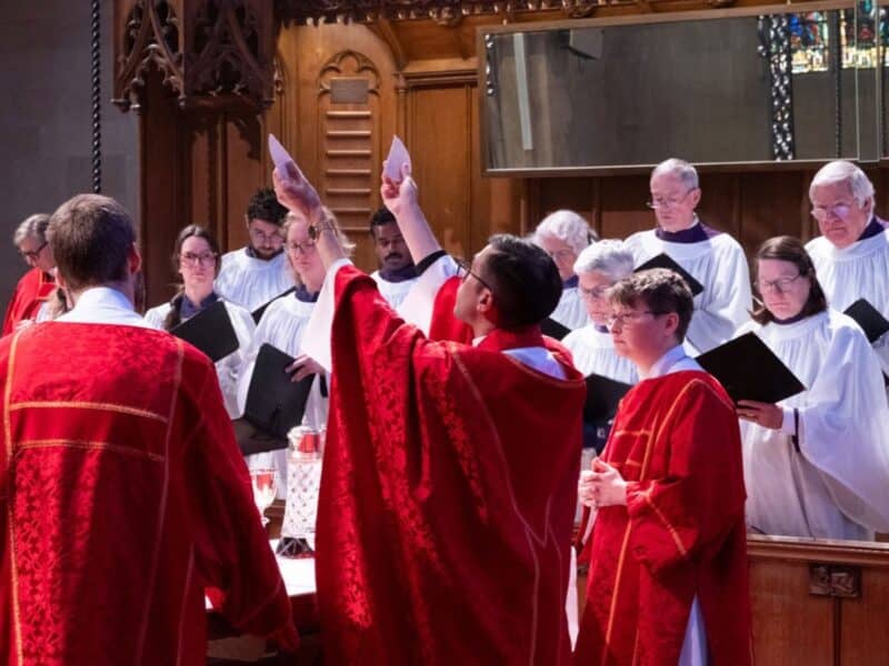 Clergy in red vestments raise their hands in prayer at the altar while the cathedral choir stands behind them.