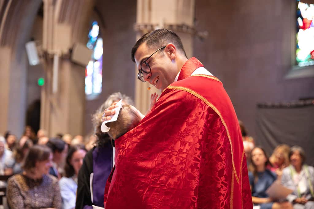 A priest holds and blesses an infant during a baptism service at St. John’s Cathedral in Denver, with the congregation gathered in the background.