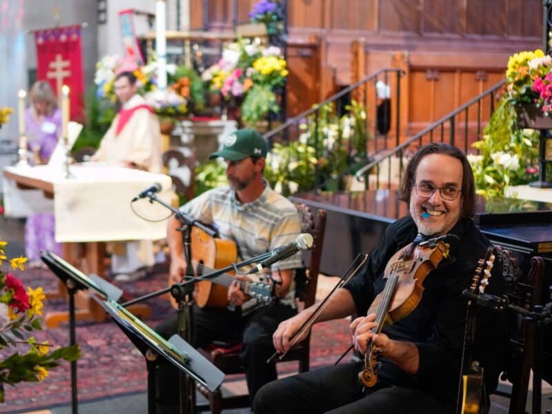 Violinist and guitarist perform during worship at St. John’s Cathedral in Denver, with clergy and flowers visible near the altar.