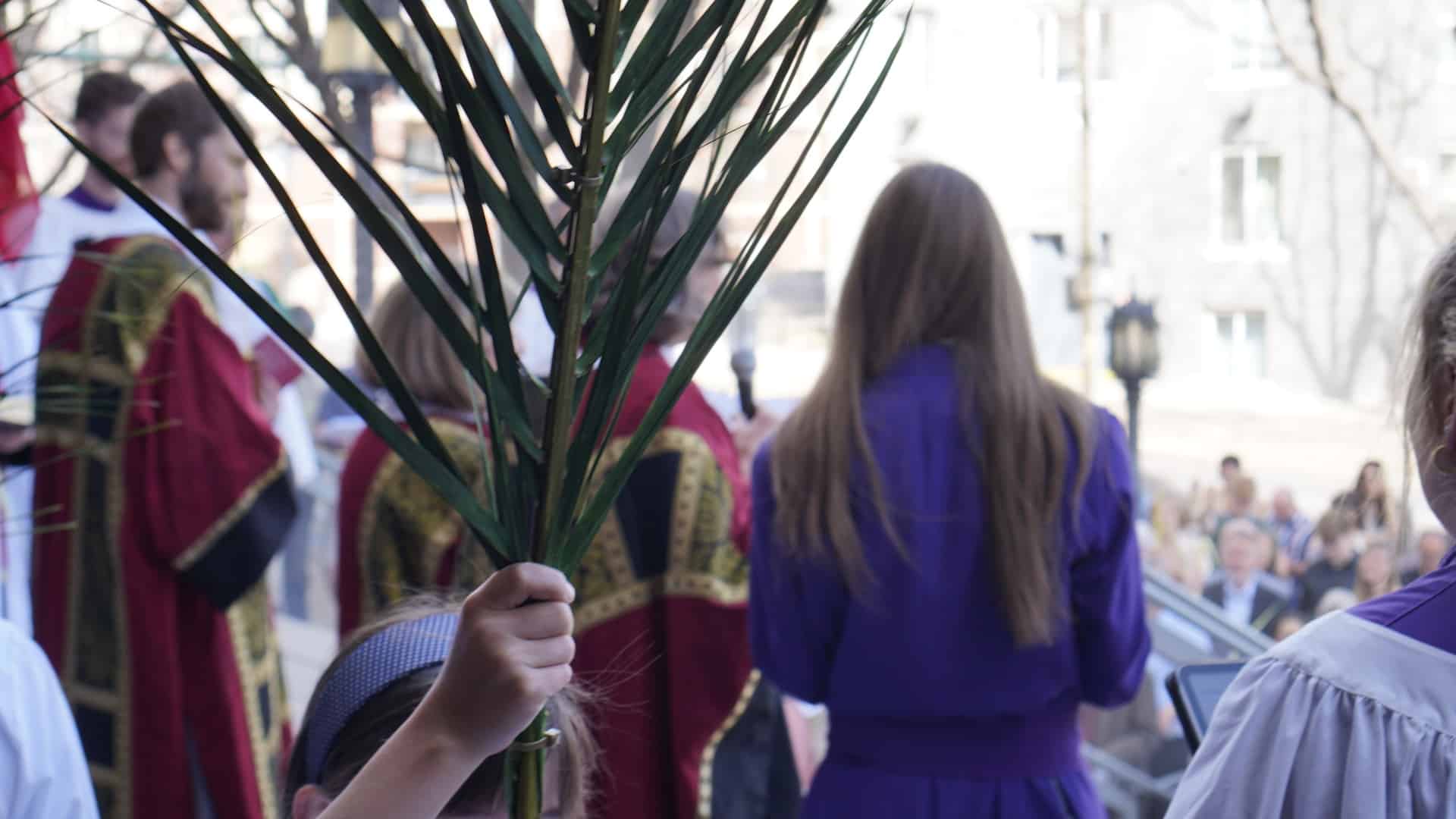 A child holds a palm branch during a Palm Sunday procession outside St. John’s Cathedral in Denver, with clergy and worshippers gathered nearby.