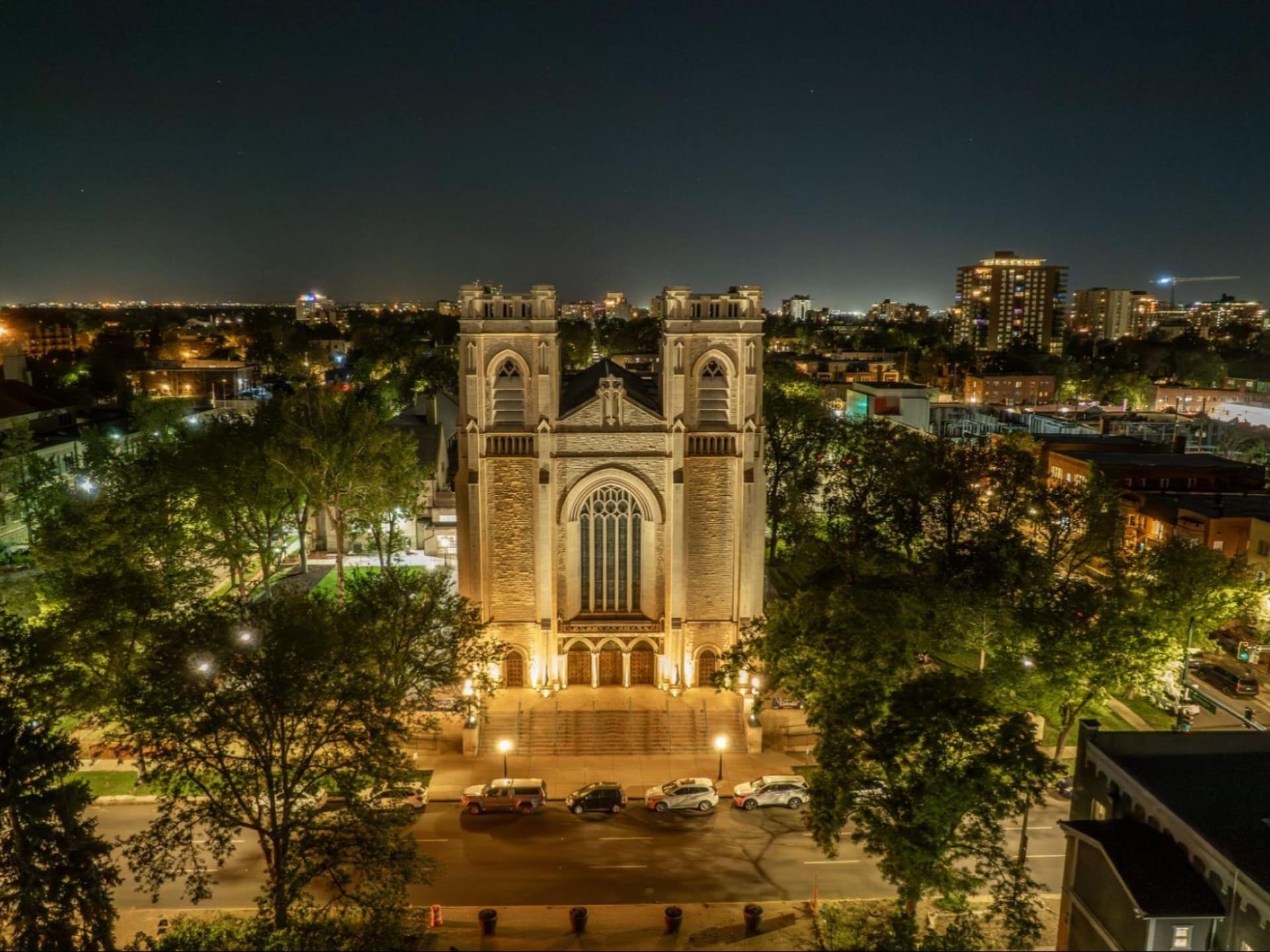 Nighttime aerial view of St. John’s Cathedral in Denver, illuminated against the surrounding city neighborhood.
