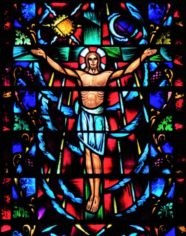 Colorful stained glass window at St. John’s Cathedral in Denver depicting Jesus with outstretched arms surrounded by vivid red, blue, green and gold glass.