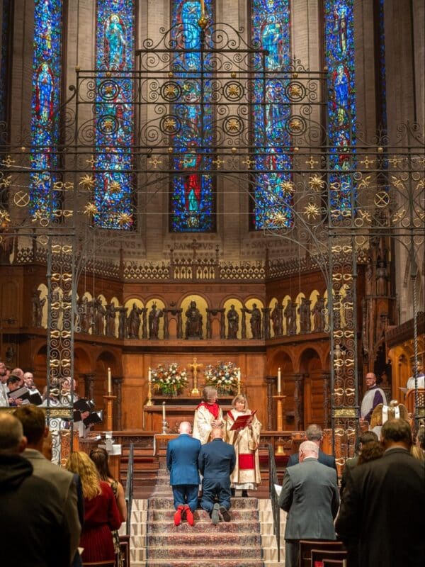 A wedding ceremony takes place inside St. John’s Cathedral in Denver beneath ornate ironwork and colorful stained glass windows.