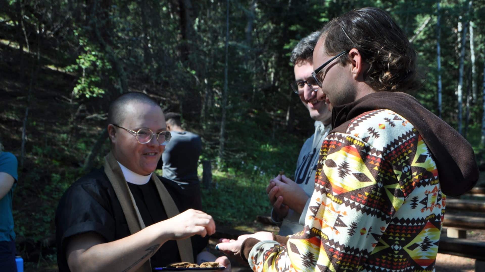 A clergy member serves communion to young adults during an outdoor gathering in a wooded setting.